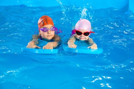 Two little girls learning how to swim in swimming pool and having fun using flutter boards