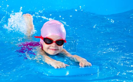 Photo for Preschool cute girl learning to swim in indoor pool with flutterboard - Royalty Free Image