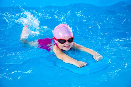 Petite fille souriante apprenant à nager dans la piscine intérieure avec planche flottante pendant les cours de natation