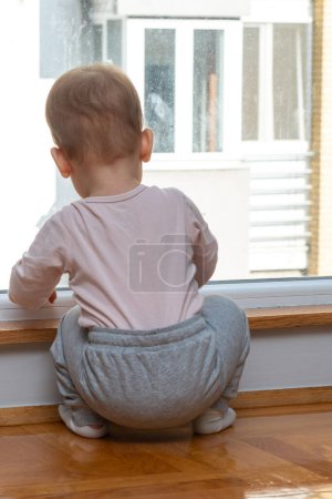 A toddler in a house looks out the window at a nearby building, dreaming of going outside for fun. Concept of a kid wanting to leave home for a walk