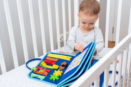Bebé de un año sentado en una cuna jugando con montessori libro ocupado