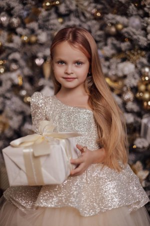 Téléchargez les photos : Photo de Noël de belle fille mignonne aux cheveux blonds dans une robe élégante posant dans un studio décoré avec arbre de Noël et cadeaux - en image libre de droit