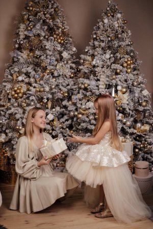 Téléchargez les photos : Photo de mode de belle famille, mère et sa fille aux cheveux blonds dans des robes élégantes posant dans un studio décoré avec arbre de Noël et cadeaux - en image libre de droit