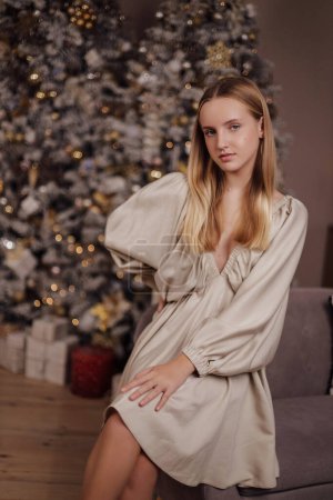Téléchargez les photos : Photo de Noël de belle fille aux cheveux blonds dans une robe élégante posant dans un studio décoré confortable avec arbre de Noël et cadeaux - en image libre de droit