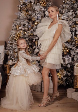 Foto de Foto de moda de hermosa familia, madre y su hija con el pelo rubio en vestidos elegantes posando en estudio decorado con árbol de Navidad y regalos - Imagen libre de derechos