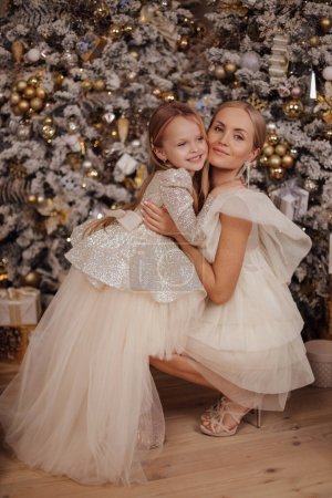 Téléchargez les photos : Photo de mode de belle famille, mère et sa fille aux cheveux blonds dans des robes élégantes posant dans un studio décoré avec arbre de Noël et cadeaux - en image libre de droit