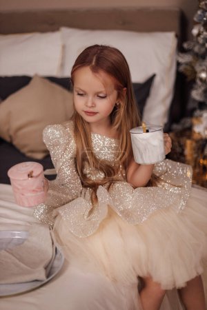 Téléchargez les photos : Photo de Noël de belle fille mignonne aux cheveux blonds dans une robe élégante posant dans un studio décoré avec arbre de Noël et cadeaux - en image libre de droit