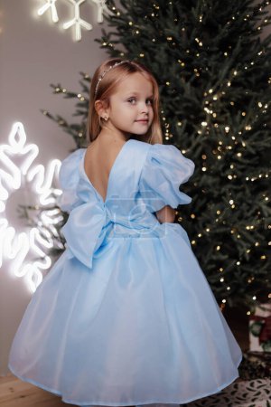 Foto de Foto de Navidad de la hermosa linda chica con el pelo rubio en vestido elegante posando en estudio decorado con árbol de Navidad y regalos - Imagen libre de derechos