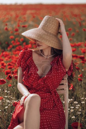 Téléchargez les photos : Photo de mode en plein air de belle femme aux cheveux foncés dans une élégante robe rouge et chapeau de paille posant dans un champ de pavot en fleurs - en image libre de droit
