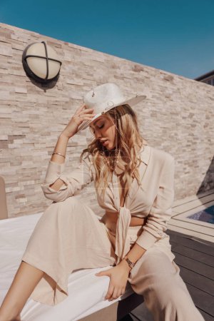 Foto de Fashion outdoor photo of beautiful woman with blond hair in elegant white clothes and hat posing in beach club near swimming pool - Imagen libre de derechos