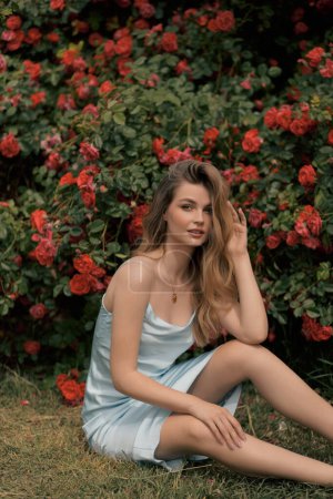 Foto de Foto de moda al aire libre de la mujer hermosa con el pelo rubio en vestido elegante posando cerca de los arbustos con rosas en flor - Imagen libre de derechos