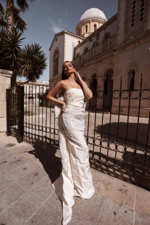 Téléchargez les photos : Mode photo extérieure de belle femme aux cheveux foncés dans une robe de mariée luxueuse avec des accessoires posant dans la rue de la ville antique - en image libre de droit