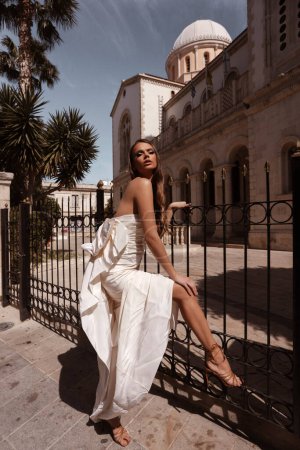 Téléchargez les photos : Mode photo extérieure de belle femme aux cheveux foncés dans une robe de mariée luxueuse avec des accessoires posant dans la rue de la ville antique - en image libre de droit