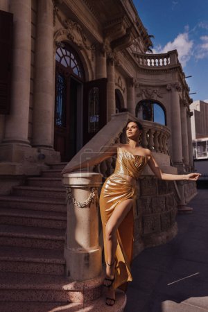 Téléchargez les photos : Photo de mode en plein air de belle femme sensuelle aux cheveux foncés dans une tenue de mode luxueuse, élégante robe dorée avec accessoires, posant devant le bâtiment dans un style classique - en image libre de droit