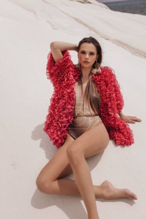 Téléchargez les photos : Photo de mode en plein air de belle femme sexy aux cheveux foncés en tenue de luxe, maillot de bain et veste de mode rouge posant sur le rocher blanc - en image libre de droit