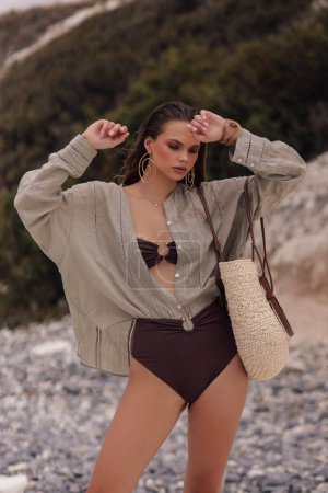 Téléchargez les photos : Photo de mode en plein air de belle femme sexy aux cheveux foncés dans un élégant maillot de bain et accessoires posant sur la plage - en image libre de droit