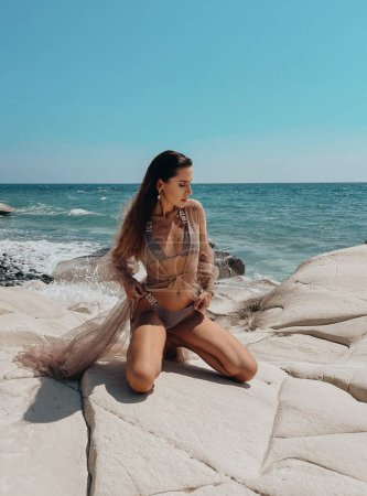 Téléchargez les photos : Photo de mode en plein air de belle femme aux cheveux foncés dans un élégant maillot de bain posant sur la plage rocheuse - en image libre de droit
