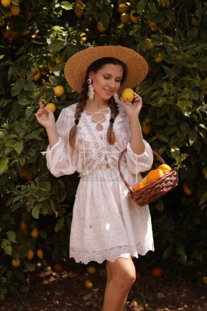 Téléchargez les photos : Photo de mode en plein air de belle femme aux cheveux foncés dans une élégante robe blanche et chapeau de paille posant dans le jardin d'été avec panier de fruits - en image libre de droit