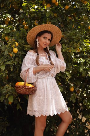 Photo for Fashion outdoor photo of beautiful woman with dark hair in elegant white dress and straw hat posing in summer garden with fruits basket - Royalty Free Image