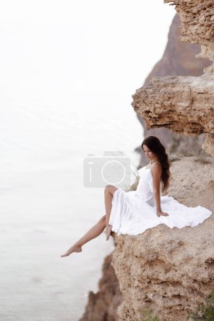 Foto de Foto de viaje de moda de mujer hermosa con el pelo oscuro en elegante vestido blanco posando en increíble lugar de la naturaleza, paisaje con montañas y el mar - Imagen libre de derechos