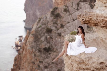 Foto de Foto de viaje de moda de mujer hermosa con el pelo oscuro en elegante vestido blanco posando en increíble lugar de la naturaleza, paisaje con montañas y el mar - Imagen libre de derechos