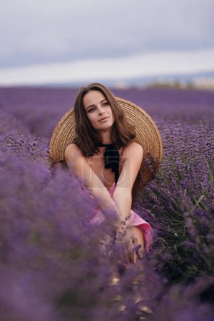 Téléchargez les photos : Photo de mode en plein air de belle femme aux cheveux foncés en robe luxueuse et grand chapeau de paille posant dans le champ de lavande en fleurs en Provence - en image libre de droit