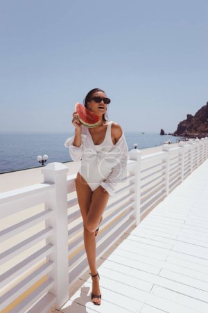 Téléchargez les photos : Photo extérieure de mode de belle femme aux cheveux foncés en élégant maillot de bain blanc et lunettes de soleil mangeant pastèque sur terrasse avec vue sur la mer - en image libre de droit