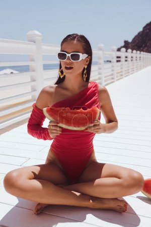 Foto de Foto al aire libre de moda de una hermosa mujer con cabello oscuro en elegante traje de baño rosa y gafas de sol comiendo sandía en la terraza con vista al mar - Imagen libre de derechos