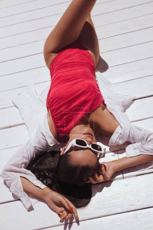 Téléchargez les photos : Photo de mode en plein air de belle femme aux cheveux foncés en élégant maillot de bain rose et lunettes de soleil mangeant pastèque sur terrasse avec vue sur la mer - en image libre de droit