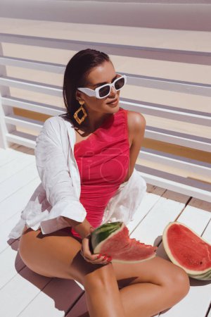 Téléchargez les photos : Photo de mode en plein air de belle femme aux cheveux foncés en élégant maillot de bain rose et lunettes de soleil mangeant pastèque sur terrasse avec vue sur la mer - en image libre de droit