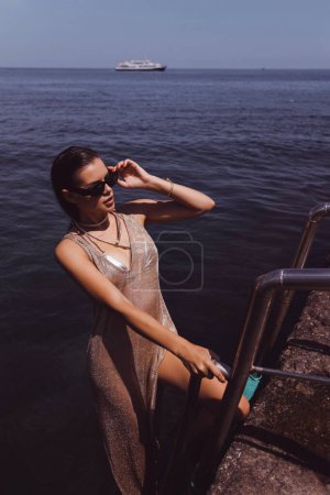 Téléchargez les photos : Mode photo en plein air de belle femme aux cheveux foncés dans des vêtements de bain élégants et lunettes de soleil nageant dans la mer à Chypre - en image libre de droit