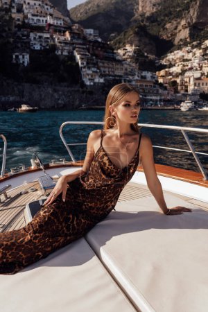 Foto de Foto de viaje de moda de hermosa mujer sexy con cabello rubio en vestido elegante relajante en el yate en Italia, ciudad de Positano en el fondo - Imagen libre de derechos