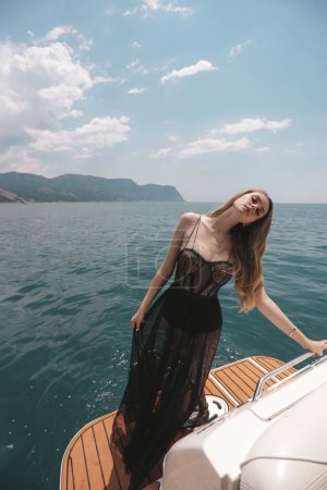 Téléchargez les photos : Photo de mode de belle femme aux cheveux blonds en robe noire luxueuse relaxant sur le yacht dans la mer Méditerranée - en image libre de droit