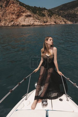 Téléchargez les photos : Photo de mode de belle femme aux cheveux blonds en robe noire luxueuse relaxant sur le yacht dans la mer Méditerranée - en image libre de droit