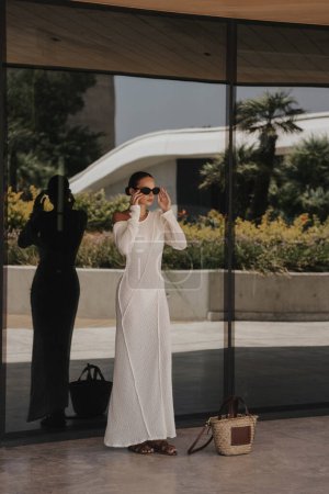 Téléchargez les photos : Photo extérieure de mode de belle femme aux cheveux foncés dans une élégante robe blanche et accessoires posant près de la fenêtre miroir de l'hôtel - en image libre de droit