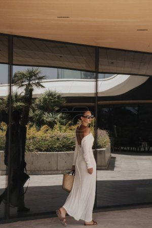 Foto de Foto de moda al aire libre de la hermosa mujer con el pelo oscuro en elegante vestido blanco y accesorios posando cerca de la ventana del espejo del hotel - Imagen libre de derechos