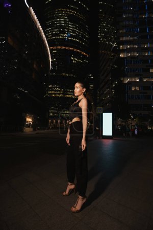 Téléchargez les photos : Mode photo extérieure de belle femme aux cheveux foncés dans une robe élégante et accessoires promenades autour de la ville de nuit avec des gratte-ciel en arrière-plan - en image libre de droit