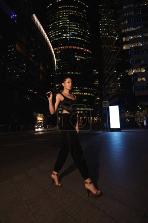 Photo for Fashion outdoor photo of beautiful woman with dark hair in elegant dress and accessories walks around the night city with skyscrapers in the background - Royalty Free Image