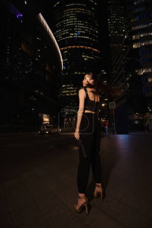Foto de Foto de moda al aire libre de la hermosa mujer con el pelo oscuro en vestido elegante y accesorios paseos por la ciudad de la noche con rascacielos en el fondo - Imagen libre de derechos