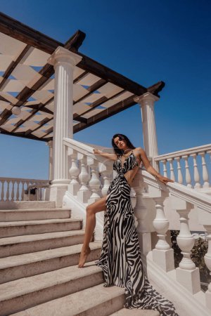 Foto de Foto de moda al aire libre de la hermosa mujer con el pelo oscuro en vestido elegante con estampado de cebra posando en el club de playa - Imagen libre de derechos