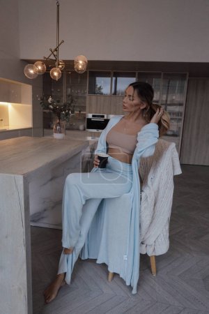Foto de Foto de moda de la mujer hermosa con el pelo rubio en ropa de casa acogedora elegante, beber café en la cocina - Imagen libre de derechos