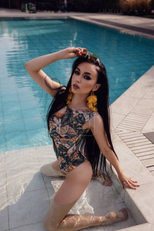 Foto de Foto de moda al aire libre de la hermosa mujer con el pelo oscuro en traje de baño elegante relajante cerca de la piscina en el club de playa - Imagen libre de derechos