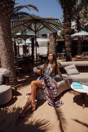 Foto de Foto al aire libre de moda de mujer hermosa con cabello oscuro en vestido de lujo y accesorios posando en club de playa al aire libre - Imagen libre de derechos
