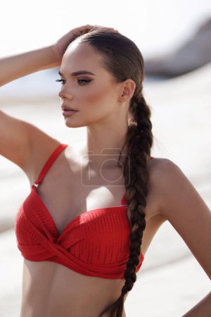 Téléchargez les photos : Photo de mode en plein air de belle femme aux cheveux foncés dans un élégant maillot de bain rouge posant sur la plage de pierres blanches à Chypre - en image libre de droit