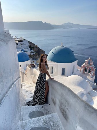 Foto de Foto de moda al aire libre de la hermosa mujer con el pelo oscuro en ropa elegante con accesorio que viaja por la isla de Santorini, iglesia azul en el fondo - Imagen libre de derechos