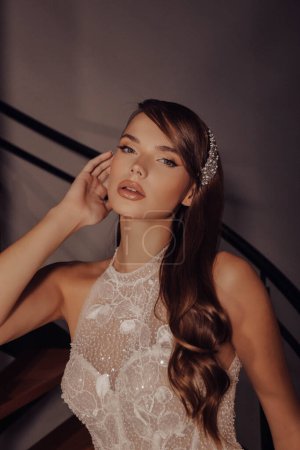 Téléchargez les photos : Photo d'intérieur de mode de belle femme aux cheveux foncés dans la robe de dentelle de mariage luxueuse posant sur les escaliers - en image libre de droit