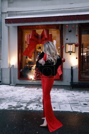 Téléchargez les photos : Photo extérieure de mode de belle femme aux cheveux blonds en manteau de fourrure élégant et robe rouge avec accessoires posant près de la vitrine décorée pour Noël - en image libre de droit