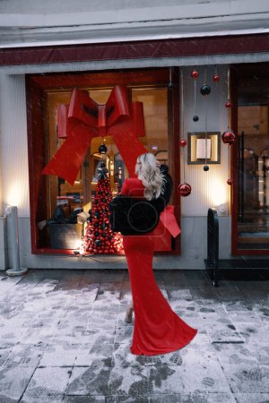 Téléchargez les photos : Photo extérieure de mode de belle femme aux cheveux blonds en manteau de fourrure élégant et robe rouge avec accessoires posant près de la vitrine décorée pour Noël - en image libre de droit