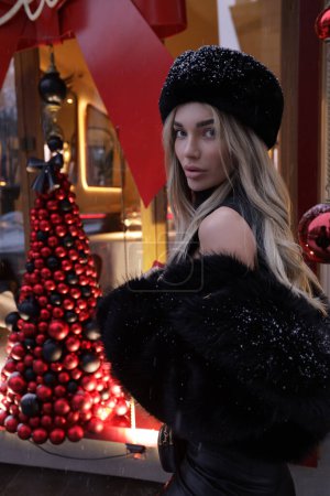 Téléchargez les photos : Photo extérieure de mode de belle femme aux cheveux blonds en manteau de fourrure élégant et accessoires posant près de la vitrine décorée pour Noël - en image libre de droit