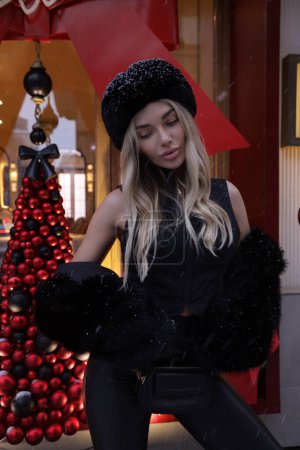 Téléchargez les photos : Photo extérieure de mode de belle femme aux cheveux blonds en manteau de fourrure élégant et accessoires posant près de la vitrine décorée pour Noël - en image libre de droit